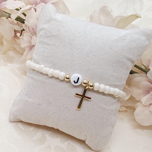 Könnte beinhalten: Ein weißes Perlenarmband mit einem goldenen Kreuzanhänger und einer weißen Perle mit dem Buchstaben "J". Das Armband wird auf einem weißen Kissen mit rosa Blumen im Hintergrund präsentiert.