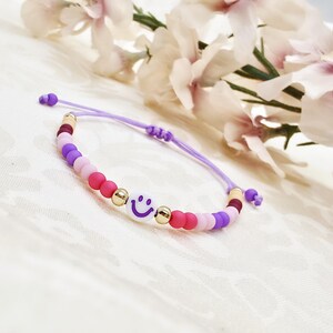 Könnte beinhalten: Ein farbenfrohes Perlenarmband mit einem Smiley-Gesicht-Design. Das Armband hat eine lila Kordel und ist mit einer Vielzahl von Perlen in Rosa-, Lila- und Goldtönen verziert. Das Smiley-Gesicht ist weiß mit einem lila Lächeln.