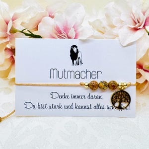 Könnte beinhalten: Goldenes Armband mit drei gesprenkelten Perlen und einem Lebensbaum-Anhänger. Das Armband wird auf einer weißen Karte mit dem Wort "Mutmacher" und inspirierendem Text präsentiert. Oben ist eine Löwensilhouette.