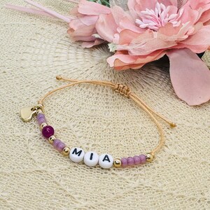 Könnte beinhalten: Ein hellbraunes Armband aus Schnur mit einem goldenen Herz-Anhänger und lila Perlen. Das Armband hat weiße Perlen mit den Buchstaben "MIA" darauf.