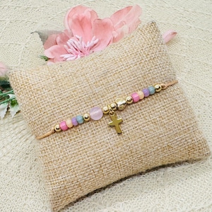 Könnte beinhalten: Ein zartes goldenes Armband mit einem Kreuzanhänger und pastellfarbenen Perlen. Das Armband liegt auf einer beigefarbenen Stoffoberfläche.