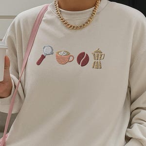 以下が含まれることがあります： クリーム色のスウェットシャツ。コーヒーをテーマにした刺繍が施されています。デザインには、ポルタフィルター、コーヒーカップ、コーヒー豆、モカポットが含まれています。ピンクのストライプのショートパンツとゴールドのチェーンネックレスを着用しています。