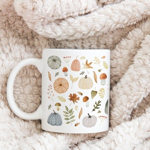 May include: A white ceramic mug with a fall-themed design. The mug features a pattern of pumpkins, leaves, mushrooms, and berries in shades of orange, brown, green, and blue.
