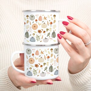 May include: Two white enamel mugs with a fall-themed design featuring pumpkins, leaves, mushrooms, and berries. The mugs are stacked on top of each other.