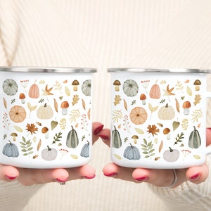 May include: Two white enamel mugs with a fall-themed design. The mugs feature a pattern of pumpkins, leaves, mushrooms, and berries in shades of orange, brown, green, and gray.