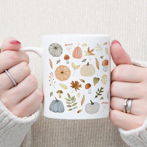 May include: White ceramic mug with a fall-themed design featuring pumpkins, leaves, and mushrooms in shades of orange, brown, green, and gray.