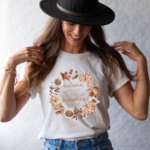 May include: White t-shirt with a floral wreath design and the text "autumn leaves and pumpkins please". A woman is wearing the shirt and a black fedora.
