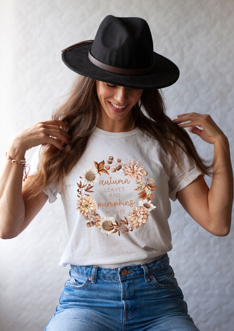 May include: White t-shirt with a floral wreath design and the text "autumn leaves and pumpkins please". A woman is wearing the shirt and a black fedora.