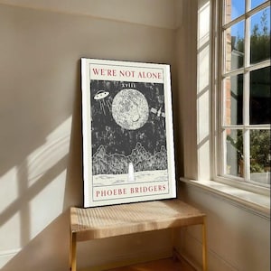 May include: A framed poster with the text "WE'RE NOT ALONE" in red, featuring a black and white illustration of a moon, UFO, and satellite. The artist's name, Phoebe Bridgers, is printed at the bottom. The poster is displayed on a woven bench.