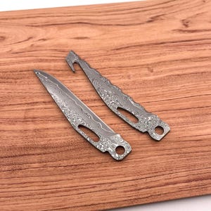 Op de afbeelding: Twee zilverkleurige mesbladen met een patroon. De bladen liggen op een houten oppervlak.