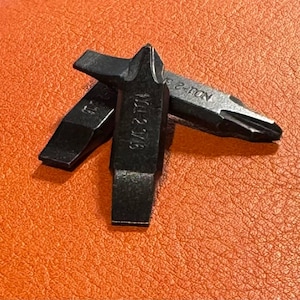 May include: Close-up of three black screwdriver bits. One bit is a flathead, and the other two are Phillips head. The bits are on an orange textured surface. The Phillips head bit has the text "No. 2-TON" and "101-2 3/16" printed on it.