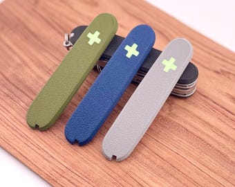 Puede incluir: Tres cubiertas protectoras de diferentes colores para una navaja suiza. Las cubiertas son verde oliva, azul marino y gris claro. Cada cubierta tiene un signo más blanco con un centro verde.