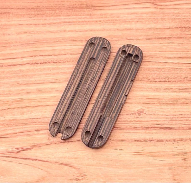 May include: Two wooden scales for a pocket knife. The scales are made of dark wood with a striped pattern. The scales have a cutout for the knife blade and a hole for the pivot pin.