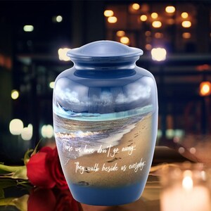 May include: Blue ceramic cremation urn with a beach scene and the quote "Those we love don't go away, they walk beside us everyday".