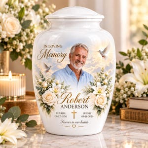 May include: A white ceramic cremation urn featuring a portrait of a man, the text In Loving Memory, Robert Anderson, Sunrise 06-12-1950, Sunset 09-18-2026, and Forever in our hearts, decorated with white roses, lilies, and two flying white doves.