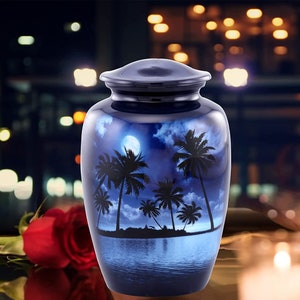 Puede incluir: Una urna de cremaci&oacute;n de metal azul y plateado con un dise&ntilde;o de palmeras y una luna llena en un cielo azul oscuro. La urna est&aacute; sobre una mesa con una rosa roja y una vela en el fondo.
