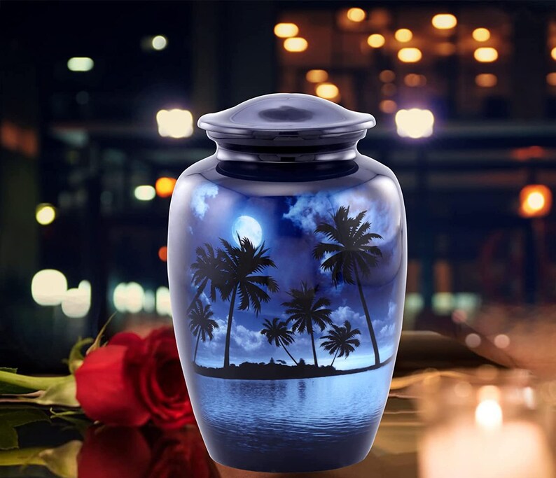 Puede incluir: Una urna de cremaci&oacute;n de metal azul y plateado con un dise&ntilde;o de palmeras y una luna llena en un cielo azul oscuro. La urna est&aacute; sobre una mesa con una rosa roja y una vela en el fondo.