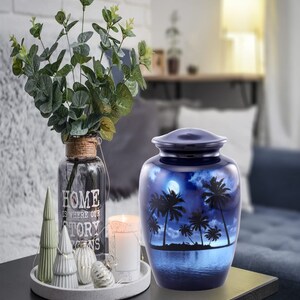 Puede incluir: Una urna de cremaci&oacute;n de cer&aacute;mica azul con una escena de palmeras y luna. La urna est&aacute; sobre una mesa de madera oscura con una vela blanca y un jarr&oacute;n de vegetaci&oacute;n.
