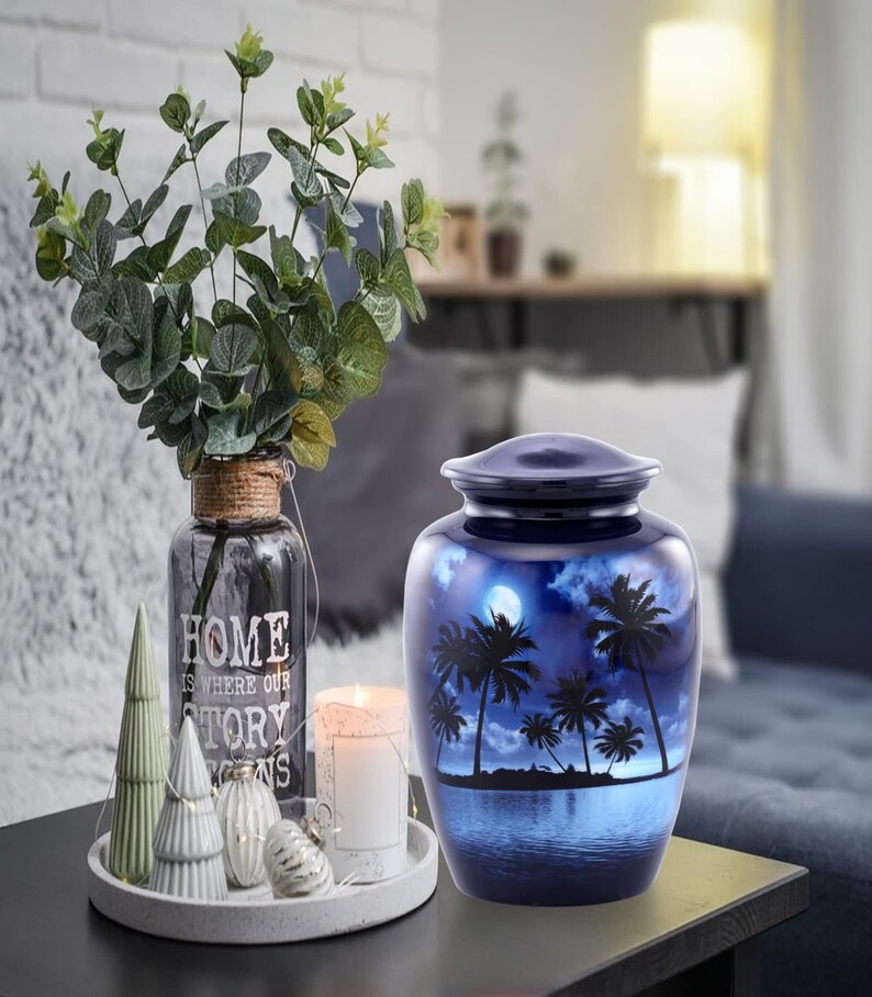 Puede incluir: Una urna de cremaci&oacute;n de cer&aacute;mica azul con una escena de palmeras y luna. La urna est&aacute; sobre una mesa de madera oscura con una vela blanca y un jarr&oacute;n de vegetaci&oacute;n.