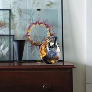 May include: A ceramic urn with a blue, orange, and yellow abstract design. The urn has a heart-shaped opening on the side. The urn is sitting on a wooden surface.