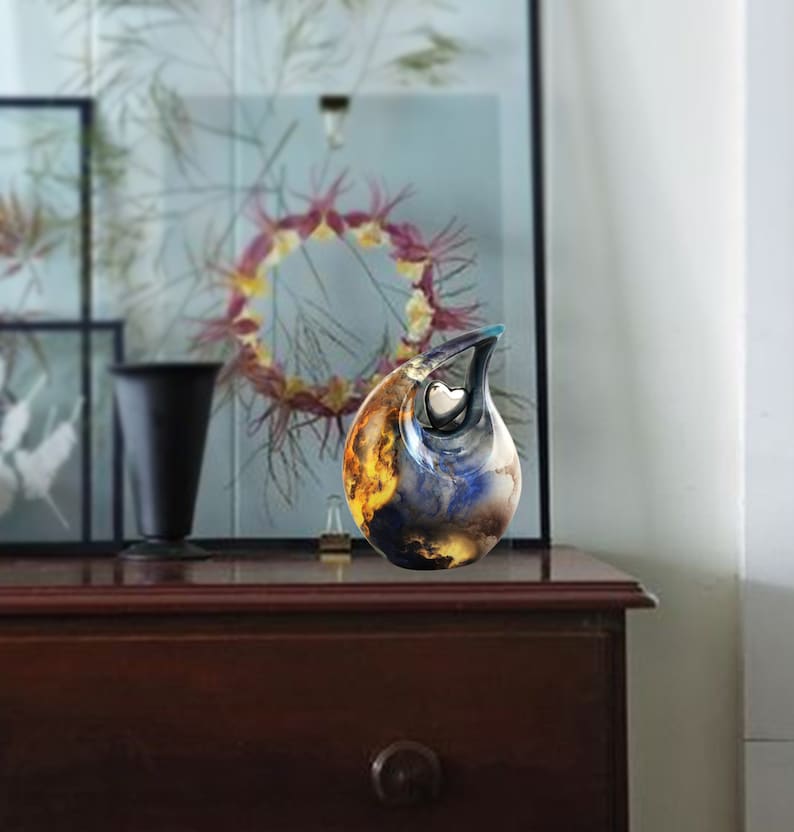 May include: A ceramic urn with a blue, orange, and yellow abstract design. The urn has a heart-shaped opening on the side. The urn is sitting on a wooden surface.