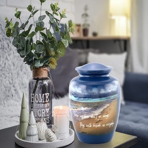 May include: Blue ceramic cremation urn with a beach scene and the quote "Those we love don't go away, they walk beside us everyday".