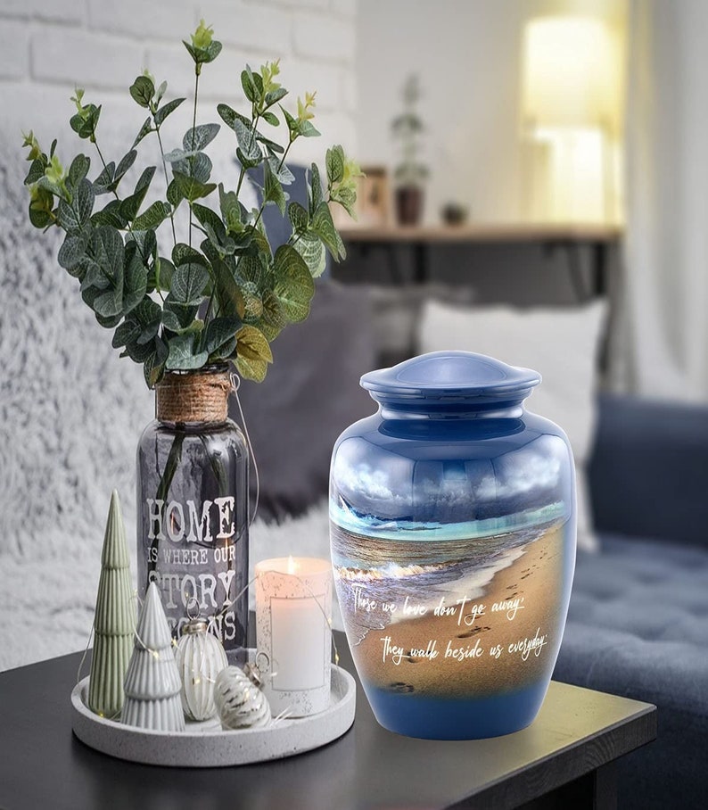 May include: Blue ceramic cremation urn with a beach scene and the quote "Those we love don't go away, they walk beside us everyday".