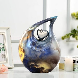 May include: A blue and yellow ceramic urn with a silver heart cutout. The urn has a swirling, abstract design.