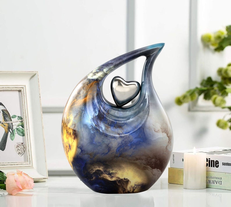 May include: A blue and yellow ceramic urn with a silver heart cutout. The urn has a swirling, abstract design.