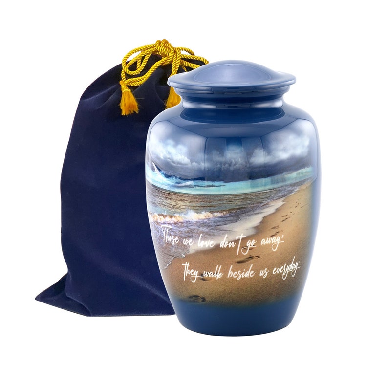May include: Blue cremation urn with a beach scene and the quote "Those we love don't go away, they walk beside us everyday". The urn comes with a blue velvet bag.