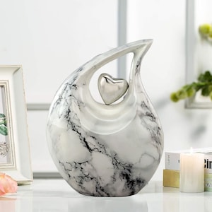 May include: White ceramic sculpture with a marble-like finish, shaped like a teardrop with a cutout in the center. A silver heart is placed inside the cutout.