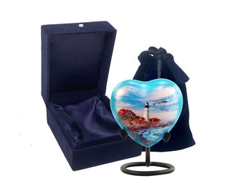 Puede incluir: Una urna de cremación de vidrio azul en forma de corazón con una imagen de un faro y un paisaje marítimo. La urna está sobre un soporte de metal negro y viene con una bolsa y una caja de terciopelo azul.