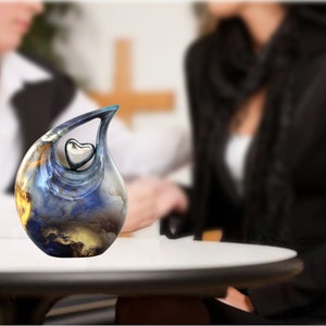 May include: A blue and gold ceramic urn shaped like a teardrop with a silver heart inside. The urn is sitting on a white table.