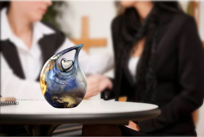 May include: A blue and gold ceramic urn shaped like a teardrop with a silver heart inside. The urn is sitting on a white table.