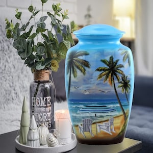 May include: A blue ceramic urn with a beach scene painted on it. The scene includes palm trees, a beach, and two chairs. The urn has a lid and is sitting on a table.
