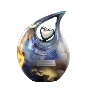 May include: A blue and gold ceramic urn with a silver heart shaped opening. The urn has a personalized plaque that reads "In Loving Memory Kevin M. Smith April 28, 1958 - October 18, 2023".
