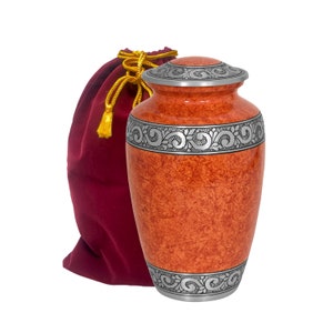 Puede incluir: Urna de cremación naranja con ribetes plateados y un diseño floral. La urna viene con una bolsa de terciopelo rojo.