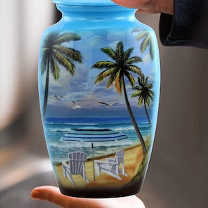 May include: A light blue ceramic urn with a beach scene painted on it. The scene includes palm trees, a beach, an umbrella, and two chairs. The urn is being held in a person's hands.