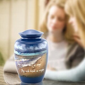 May include: Blue cremation urn with a beach scene and the words "Those we love don't go away, they walk beside us everyday".