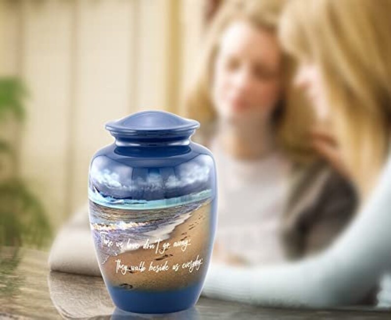 May include: Blue cremation urn with a beach scene and the words "Those we love don't go away, they walk beside us everyday".
