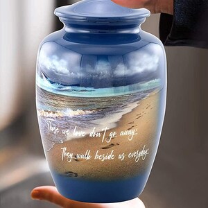 May include: Blue ceramic urn with a beach scene and the quote "Those we love don't go away, they walk beside us everyday".