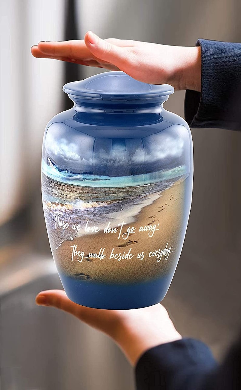 May include: Blue ceramic urn with a beach scene and the quote "Those we love don't go away, they walk beside us everyday".