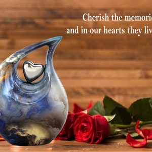 May include: A blue and gold ceramic urn with a silver heart on top. The urn is sitting on a wooden surface with red roses and petals.