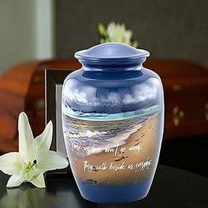 May include: Blue and gold cremation urn with a beach scene and the quote "Those we love don't go away, they walk beside us everyday".