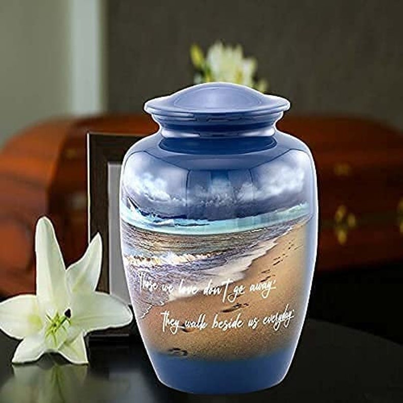 May include: Blue and gold cremation urn with a beach scene and the quote "Those we love don't go away, they walk beside us everyday".