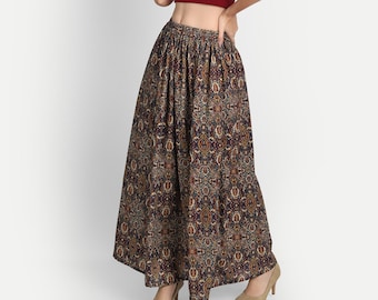 May include: A woman is wearing a maroon crop top and a long, flowing skirt with a paisley print. The skirt has a wide waistband and a flowing silhouette. The woman is wearing beige heels.