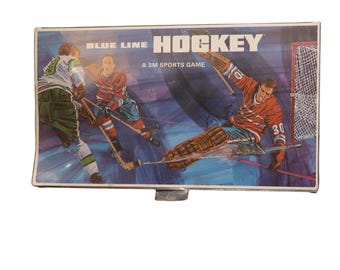 Könnte beinhalten: Vintage Blue Line Hockey Spielbox. Die Box zeigt eine farbenfrohe Illustration von Eishockeyspielern in Aktion, mit einem Torwart, der sich zum Blocken eines Schusses stürzt. Der Text "BLUE LINE HOCKEY" und "A 3M SPORTS GAME" sind sichtbar.