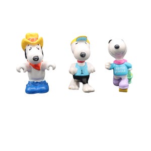 May include: Three Snoopy figurines in various outfits. One wears a cowboy hat and bandana, another a blue athletic top with the number 1, and the third a blue shirt with "Easter Beagle" text. Each figure is white with black accents.