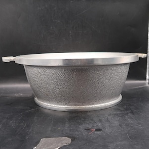 May include: A silver metal bowl with a textured surface and a smooth rim. The bowl has two small handles on either side and sits on a flat base. The bowl is likely for cooking or serving.