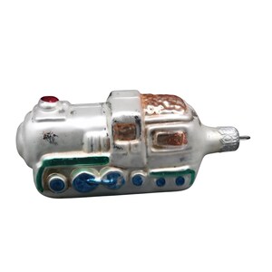 May include: A vintage Christmas ornament shaped like a silver train engine. The ornament has a red light on top, brown detailing, and green and blue accents along the bottom. It is designed to hang from a tree.